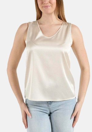 Femme portant un top en satin crème sans manches et un jean bleu clair, debout, les mains reposant près des hanches sur un fond blanc uni.