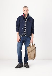 Marineblaues Bomberjackett mit einem gestreiften Hemd darunter, blauen Jeans und schwarzen Sneakers; trägt eine sandfarbene Tasche mit einem Schulterriemen und mehreren Taschen.