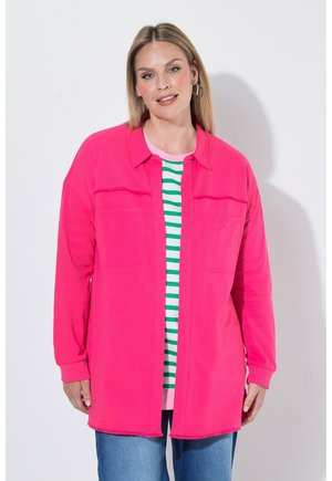 Kvinde iført en lys pink langærmet skjorte over en grøn- og hvidstribet top med blå jeans, stående mod en ensfarvet hvid baggrund.