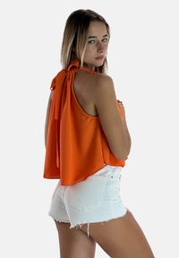 Top arancione senza maniche con design a legatura posteriore, caratterizzato da una vestibilità ampia e una consistenza morbida, abbinato a pantaloni corti in denim bianco con effetto usurato.