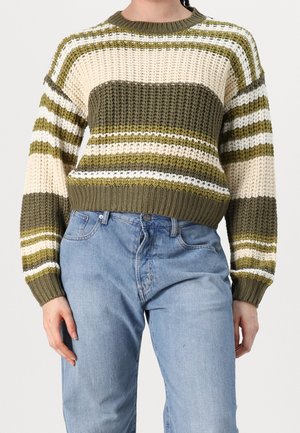 Personne portant un pull en grosse maille rayé olive, crème et blanc, avec un jean taille haute bleu clair, debout devant un fond uni.
