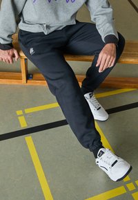 Pantalones de chándal negros con puños elásticos y bolsillos laterales, combinados con zapatillas blancas. La sudadera gris tiene texto morado y se lleva de manera casual.