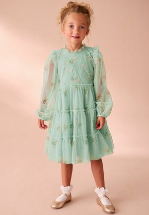 Una niña pequeña con cabello rizado lleva un vestido floral verde menta con mangas de globo transparentes, calcetas blancas con flores y zapatos Mary Jane dorados.