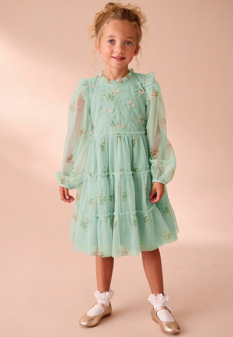 Ragazzina con capelli ricci che indossa un vestito floreale verde menta con maniche a palloncino trasparenti, calze bianche con fiori e scarpe Mary Jane dorate.
