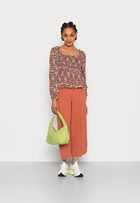 Blusa floral de mangas longas em vermelho e verde, combinada com calças largas laranja. Acessório de uma mala verde e sapatilhas brancas com detalhes em néon.