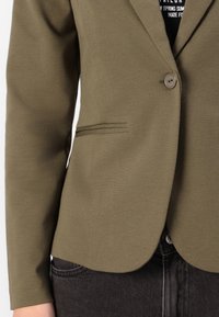 Olijfgroene blazer van een gestructureerde stof, voorzien van een enkele knoopsluiting, notch revers en borstzak, gecombineerd met donkere spijkerbroek.
