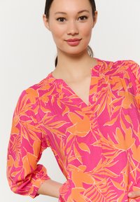 Kurzarmbluse mit V-Ausschnitt, auf einem rosa Hintergrund mit kräftigen orangenen Blumenmustern. Leichter Stoff mit leichtem Struktur.