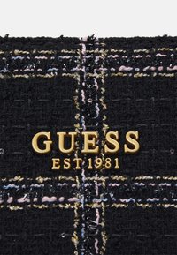 Un tissu noir texturé présente un motif tissé avec des fils dorés, roses et bleus. Un logo doré indique en évidence "GUESS EST 1981".