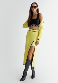 Gonna gialla in maglia con spacco laterale e accenti neri, abbinata a un crop top nero con un nastro con logo e a un cardigan giallo. Stivali neri fino al ginocchio.