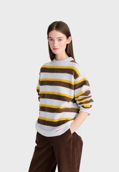 Jeune femme aux cheveux bruns et raides, portant un pull moelleux rayé marron, jaune et blanc, et un pantalon en velours marron, les mains dans les poches.