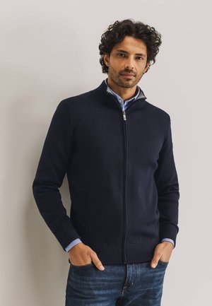 Navy zip-up sweater in a knit fabric with a ribbed hem and cuffs. Worn over a light blue collared shirt, featuring a high collar design.