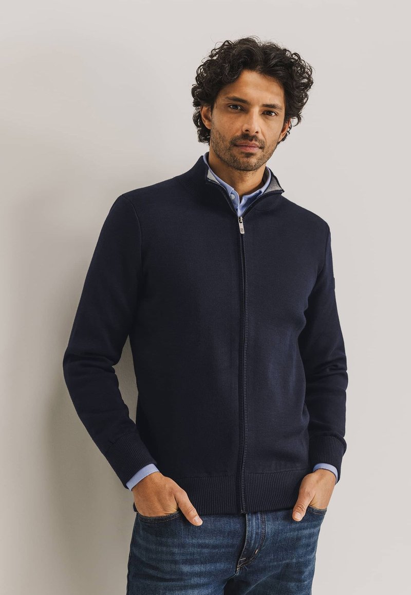 Marineblauer Zip-Up-Pullover aus Strickstoff mit geripptem Saum und Bündchen. Über einem hellblauen Hemd mit Kragen getragen, das ein hochgeschlossenes Design aufweist.