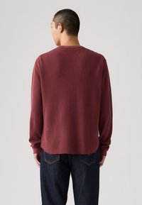 Άτομο που φοράει μακρυμάνικο, υφασμάτινο πουκάμισο μπορντό και σκούρο μπλε τζιν, με την πλάτη προς τα εμπρός σε απλό ανοιχτό φόντο.