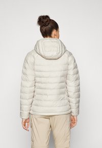 Lys beige pufferjakke med hette, quiltet tekstur, sidelommer og elastiske mansjetter. Kombinert med beige bukser, sett fra baksiden.