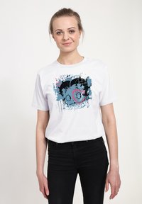 T-shirt bianco in cotone con una grafica astratta colorata con accenti blu, rosa e neri, indossato con jeans neri.