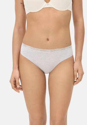 Mujer con sujetador blanco y ropa interior gris claro con encaje, de pie con los brazos relajados a los lados sobre un fondo liso.