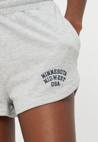 Κοντινό πλάνο γκρι σορτς με ελαστική μέση και μαύρο κείμενο «MINNESOTA MIDWEST USA» σε μηρό με καστανό δέρμα, χέρι στην τσέπη.