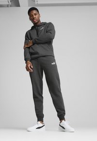 PUMA sportswear sæt i mørkegrå, der inkluderer en zip-hættejakke og tapered bukser med hvide logo accenter. Stoffet er blødt, tekstureret og afslappet.