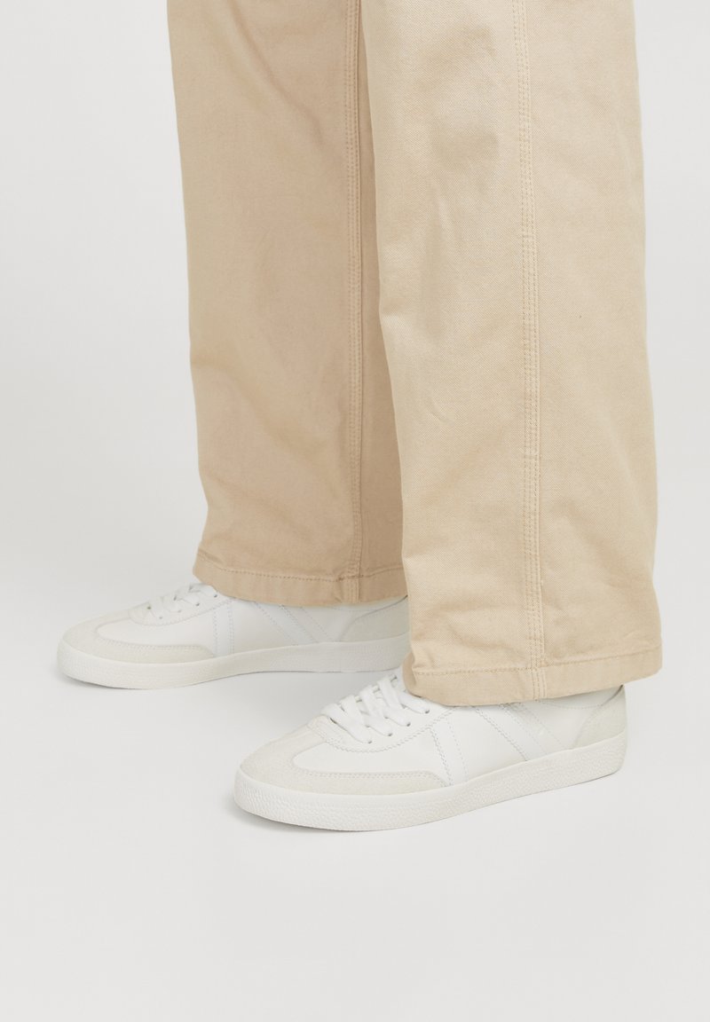 Sneakers bianche in pelle strutturata e pannelli in rete, con un design low-top e chiusura con lacci, indossati con pantaloni beige chiaro.