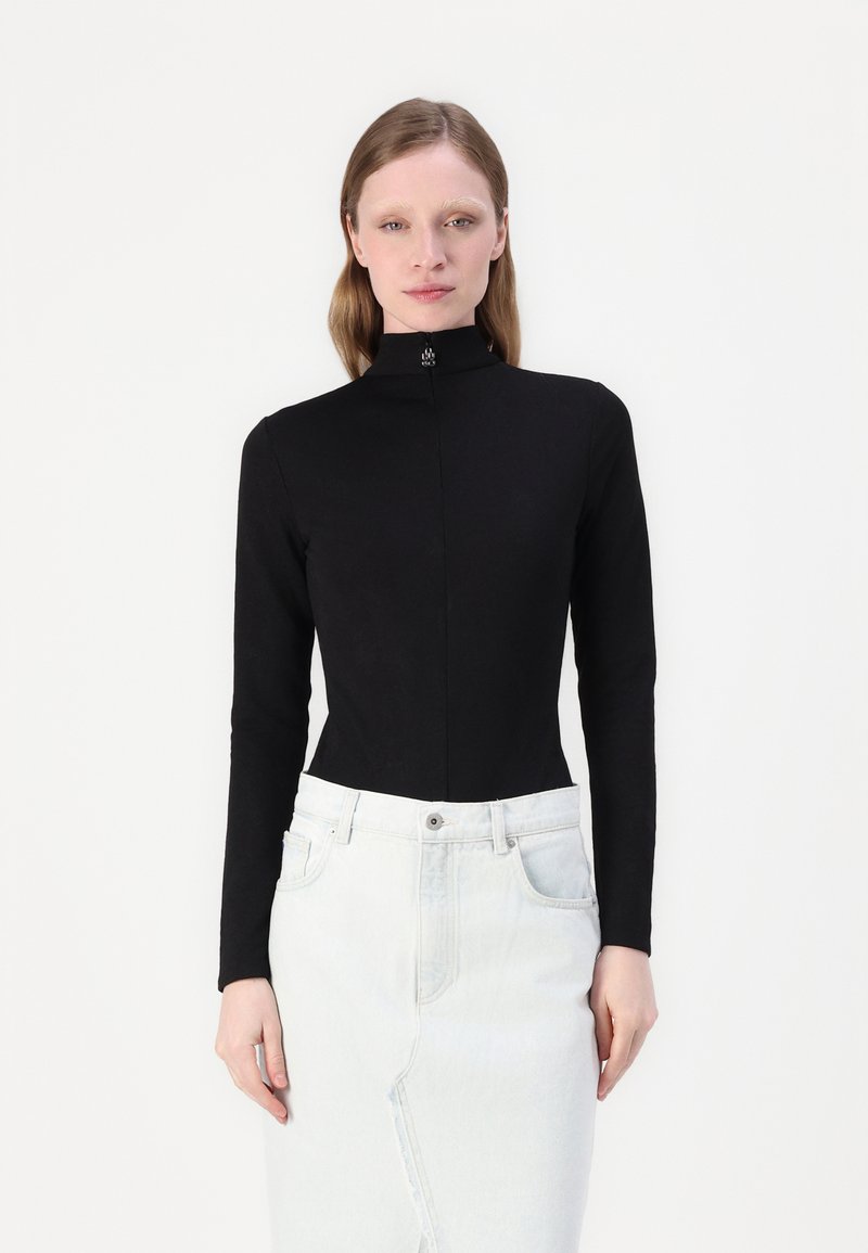 Crni dugouhi turtleneck top s uskim dizajnom, izrađen od glatkog materijala, s suptilnim logotipom na vratu. Uparen s laganom traper suknjom.