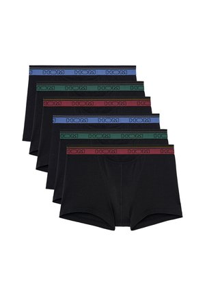 Hombre musculoso de pie descalzo, vistiendo unos bóxers negros con una cinturilla verde oscuro, mirando ligeramente hacia su derecha sobre un fondo blanco.
