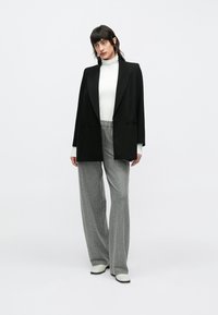 Femeie purtând un sacou negru peste un pulover alb cu guler înalt, pantaloni largi cu dungi gri și pantofi albi, stând în fața unui fundal simplu.