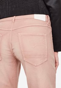 Detailní záběr na růžové džíny s zadními kapsami a bílou etiketou, nošené s texturovaným černým svetrem s dlouhým rukávem.