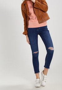 Bronze suede jacket over a pink sweatshirt, paired with blue skinny jeans featuring knee rips and cuffed hems, complemented by white sneakers.