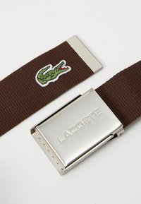 Hnedý tkaný pás s výšivkou zeleného krokodíla. Strieborný kovový spon s vygravírovaným nápisom "LACOSTE".