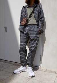 Svart mönstrad jacka och byxor med vit text, ihop med en grön polo. Vita sneakers och en brun leopardmönstrad axelväska.