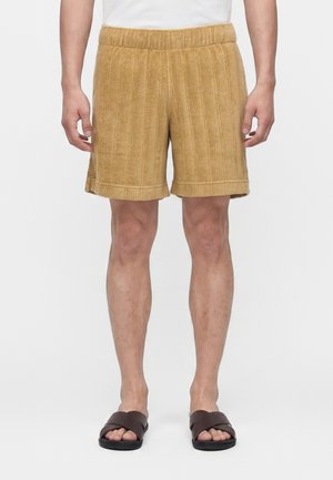 Homme portant un short en éponge côtelé beige, une chemise blanche et des sandales marron foncé à brides croisées, debout devant un fond blanc uni.