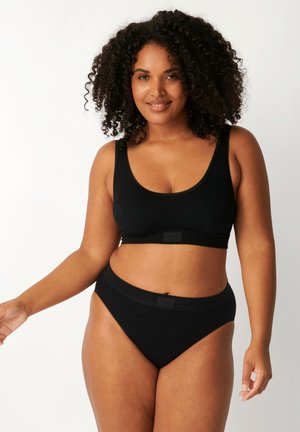 Donna dai capelli ricci che sorride, indossa un reggiseno sportivo nero e mutandine nere a vita alta, in piedi davanti a uno sfondo bianco semplice.