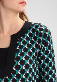 Multikleuren gebreide top met een zwarte halslijn, voorzien van een patroon van groene, blauwe en witte bloemvormen op een donkere achtergrond.