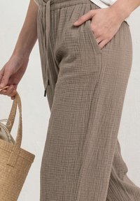 Femme portant un pantalon taupe ample et texturé avec cordon de serrage, main dans la poche, tenant un sac en paille tressée par la sangle.
