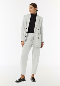 Lichtgrijs pak met een dubbelzijdige blazer en op maat gemaakte broek, gecombineerd met een zwarte coltrui en glanzende zwarte loafers.