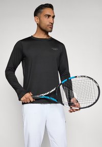 Black long-sleeve athletic top with a crew neck, featuring a small logo on the chest, paired with white athletic pants and holding a tennis racket.
