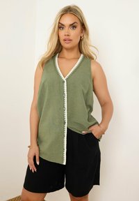 Vihreä hihaton paita V-aukolla, jossa on valkoiset yksityiskohdat ja nappi, yhdistettynä korkeavyötäröisiin mustiin shortseihin. Pehmeä kangas.