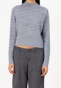 Un pull en tricot bleu clair au design court, présentant un motif texturé et des bordures côtelées au niveau du cou et des poignets. Accordé avec un pantalon gris.