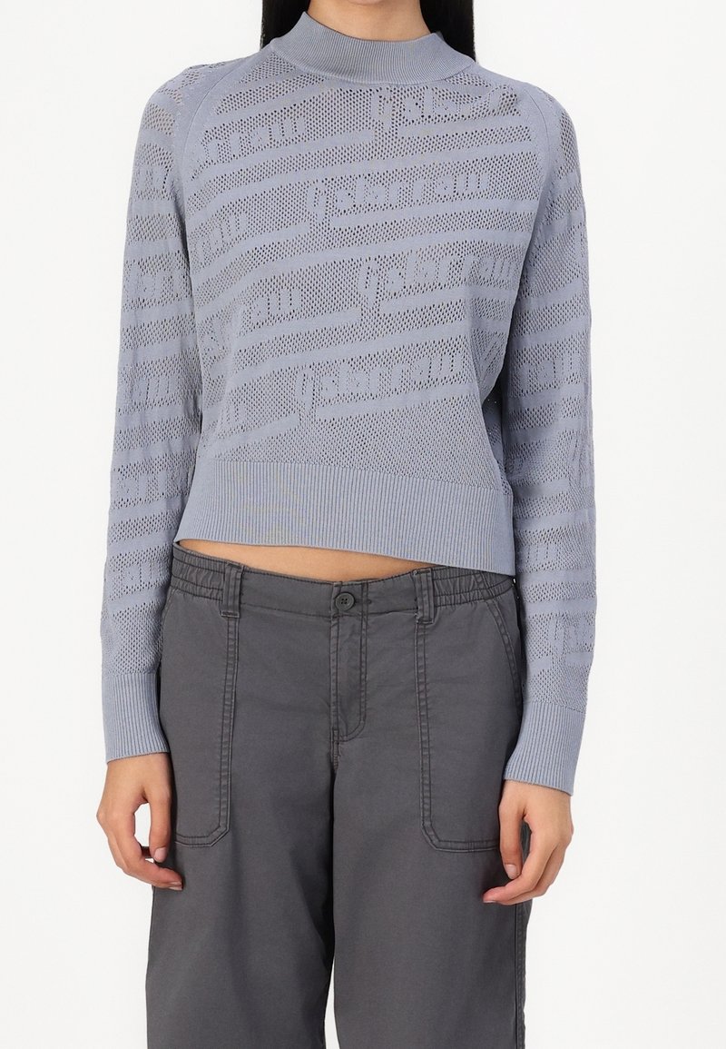 Un pull en tricot bleu clair au design court, présentant un motif texturé et des bordures côtelées au niveau du cou et des poignets. Accordé avec un pantalon gris.