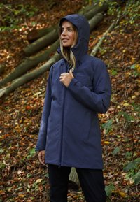 Giacca impermeabile blu navy con cappuccio aderente e chiusura con zip, caratterizzata da una texture liscia e un logo discreto sul petto, indossata all'aperto.