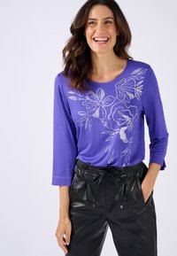 Paarse, langmouwtop met een zilveren bloemenprint. Gecombineerd met zwarte, strakke broek met een elastische tailleband en trekkoord.