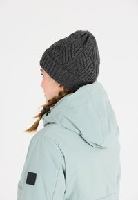 Grijze gebreide beanie met een textuurpatroon, voorzien van een geribbelde rand. Draagt een lichtblauwe jas, weergegeven in een zijprofiel van de drager.