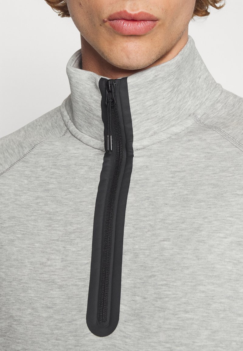 Pullover zippé gris clair avec col montant, présentant une fermeture éclair noire et des coutures contrastées, fabriqué en matière texturée.