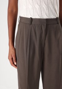 Pantalon marron sur mesure avec une texture lisse, des plis et une ceinture plate. La main du modèle repose sur la couture latérale.