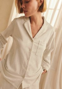 Femme portant une chemise à manches longues beige clair avec des boutons et une poche, se tenant à l'intérieur contre des rideaux beiges avec une douce lumière naturelle.