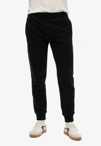 Zwarte sweatpants van zachte stof, met een elastische tailleband en ribgebreide boorden, gecombineerd met witte sneakers en zichtbare sokken.
