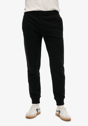 Pantalones de chándal negros hechos de una tela suave, con una cinturilla elástica y puños acanalados, combinados con zapatillas blancas y calcetines visibles.