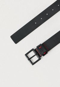 Μαύρη δερμάτινη ζώνη με τετράγωνη μαύρη αγκράφα και κόκκινες ραφές ως διακοσμητικά στοιχεία. Διαθέτει ρυθμιζόμενες τρύπες για προσαρμογή στο μέγεθος.