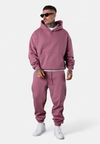 Hoodie rose et joggings assortis en tissu doux, avec une poche frontale et des poignets élastiques, accompagnés de baskets blanches.
