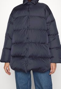 Marineblaue Steppjacke mit hohem Kragen und Druckknöpfen. Verfügt über ein gesteppte Design mit weiten Ärmeln und einer leicht oversized Passform.